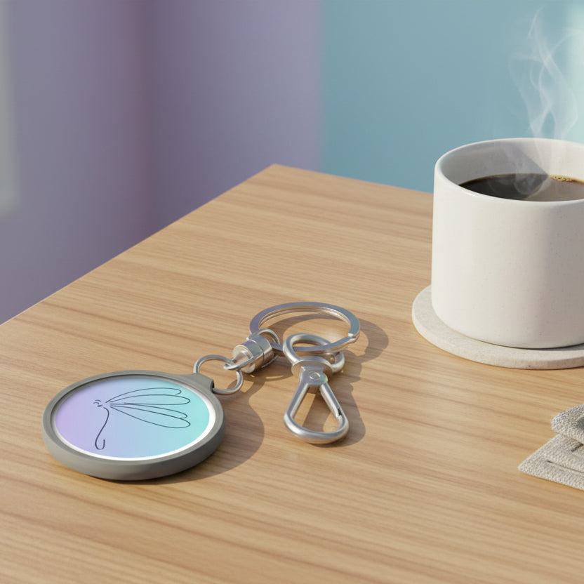Keychain with a pastel-colored circular charm featuring a dragonfly design on a coffee table with a cup of coffee next to it.