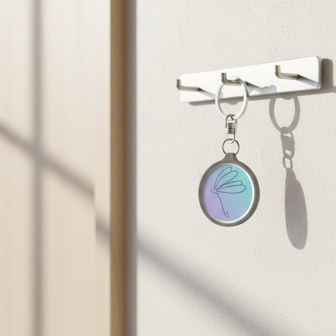 Keychain with a blue purple circular charm featuring a dragonfly stick drawing design hung on key holder by a door