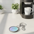 Keychain with a pastel-colored circular charm featuring a dragonfly stick drawing design on a kitchen counter with coffee machine, cut and plants next to it.