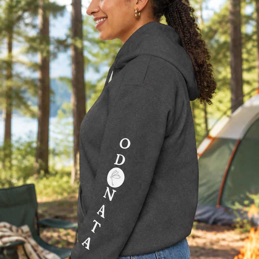 A woman shown in side profile wearing a dark charcoal gray hoodie with a white vertical 'ODONATA' graphic printed down the left sleeve, paired with blue jeans against a plain white background.