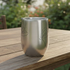 Silver tumbler with dragonfly stick drawing on it's side showing half of the drawing on the other side sitting on a rustic outdoor dining table