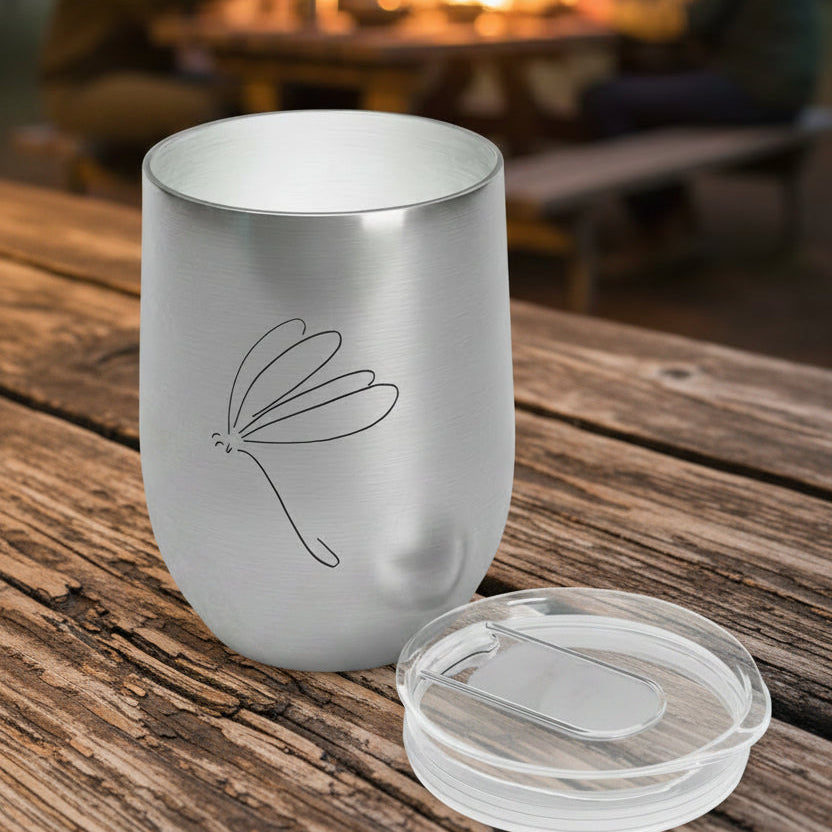 Silver tumbler with a lid that has a dragonfly stick drawing on a picnic table with people gathered around a fire in the background