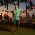 Man wearing a hawaiian shirt with a dragonfly on a leaf design at a luau with torches and other people in the background