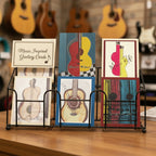 Five greeting cards featuring artistic guitar illustrations, arranged on a card rack in a music store with guitars hung in the background