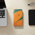 A smartphone wallet case with an abstract orange, green, and yellow design is displayed on a light wood desk alongside a laptop, a pen, and a black tablet.