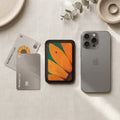 A slim rectangular card holder or wallet with a black stitched border, featuring a close-up floral print of orange petals and green leaves with a ladybug on a table with a phone and 2 credit cards next to it.