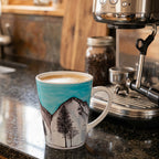 A white ceramic mug featuring a hand-painted Yosemite winter mountain landscape with a bright turquoise sky, snow-covered peaks, and dark pine trees, shown on a clean white background.