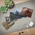 A styled flat-lay of a gray desk mat on a wooden table, featuring printed landscape photos of us parks, a silver stylus, a white wireless earbud case, earbuds, binder clips, and two green succulent plants.