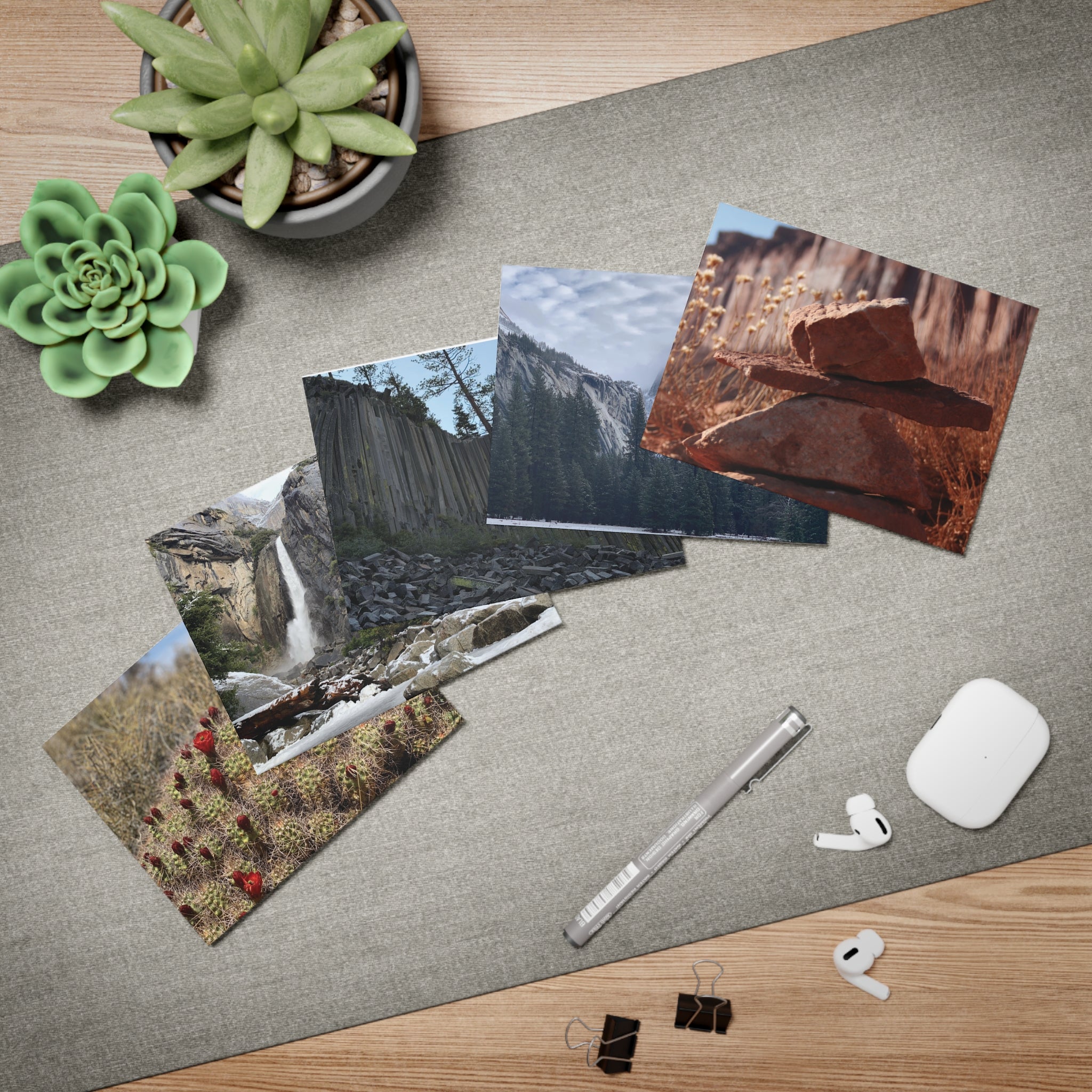 A styled flat-lay of a gray desk mat on a wooden table, featuring printed landscape photos of us parks, a silver stylus, a white wireless earbud case, earbuds, binder clips, and two green succulent plants.