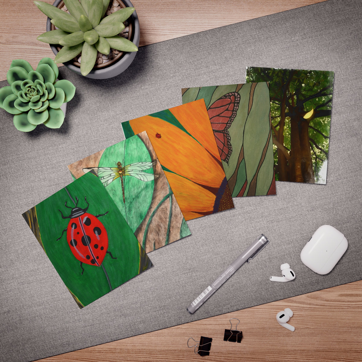 A styled flat-lay of five colorful art prints featuring nature themes (ladybug, dragonfly, butterfly, trees, leaves) arranged on a gray desk mat with a silver pen, white earbuds, clips, and potted succulents.