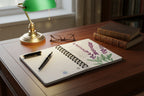 Open notebook with lavender plant design on desk with lamp, glasses, book and a pen