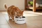 White pet bowl with 'pet name here' text on kitchen floor with a cat drinking water from it.