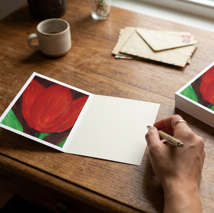 Red Tulip Greeting card on desk with a hand about to write something and with a coffee, letters and a stack of the cards next to it