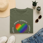 Folded green t-shirt with rainbow heart and 'Spread Kindness' text, surrounded by casual items on a light surface.