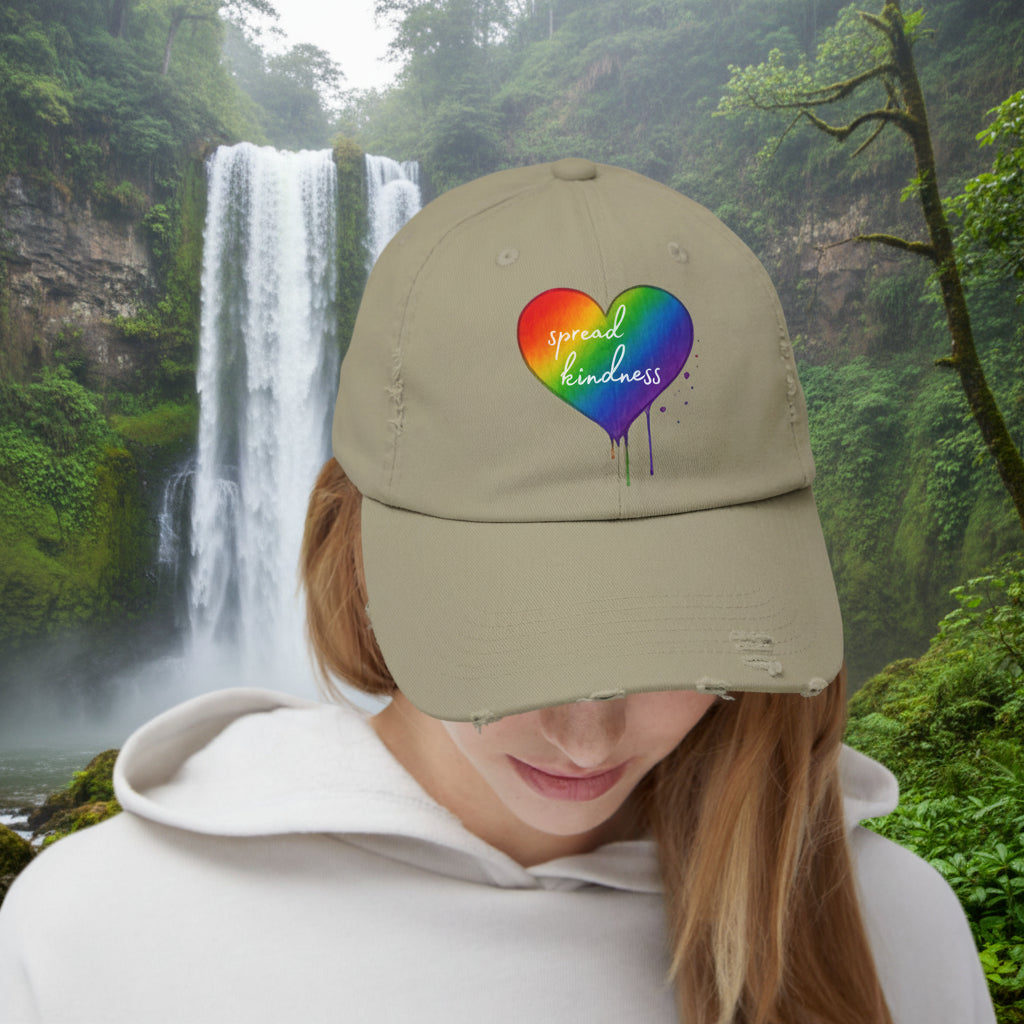 Person wearing a beige cap with a rainbow heart design and 'spread kindness' text with waterfall behind.