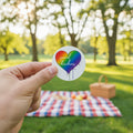 Rainbow heart with 'spread kindness' text held by a hand at at park setting.