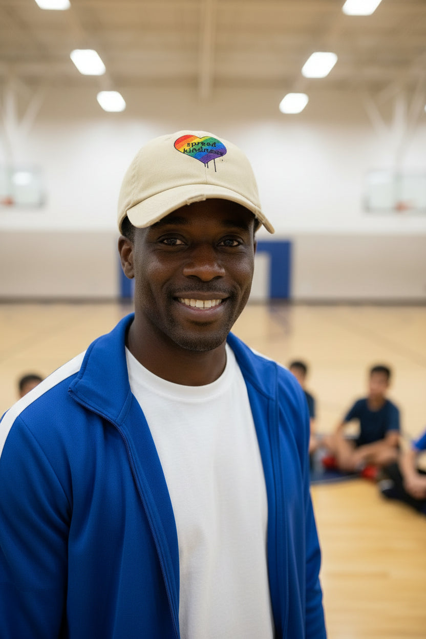 Beige cap with a rainbow heart and 'spread kindness' text on a male gym coach at a school basketball gym