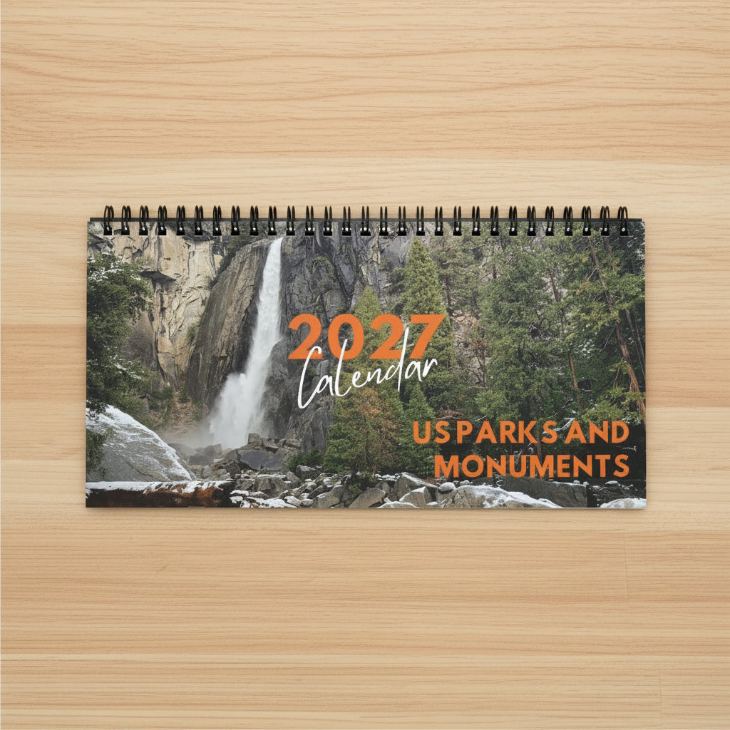 A spiral-bound 2027 wall calendar titled 'US Parks and Monuments' featuring a scenic photograph of a waterfall in a rocky, forested landscape on the cover, on top a light wood desk