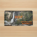 A spiral-bound 2027 wall calendar titled 'US Parks and Monuments' featuring a scenic photograph of a waterfall in a rocky, forested landscape on the cover, on top a light wood desk