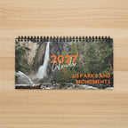 A spiral-bound 2027 wall calendar titled 'US Parks and Monuments' featuring a scenic photograph of a waterfall in a rocky, forested landscape on the cover, on top a light wood desk