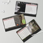 Three spiral-bound desk calendars with nature-themed photographic backgrounds are arranged on a light gray surface with binder clips and a white pen nearby.