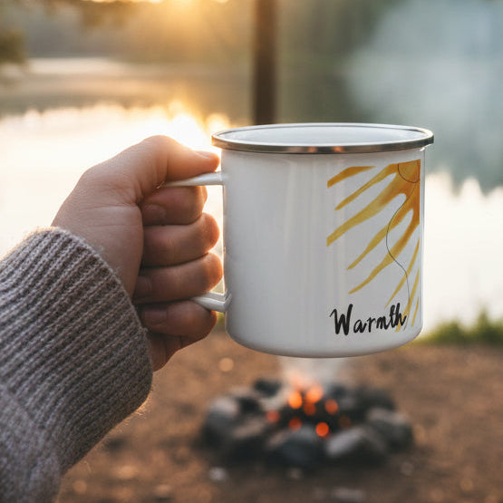 White mug with sun design and 'Warmth' text held by a hand at a campsite