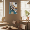 An abstract canvas wall art print featuring a stylized coffee cup and circular patterns in blue, beige, brown, black, and white, displayed on a warm beige wall in a cozy, sunlit cafe interior with wooden tables and chairs.
