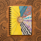 A spiral-bound notebook with a colorful abstract cover design featuring a large yellow sunflower-like shape, blue patterned sections, a red polka-dot area, black-and-white stripes, and purple accents, shown on a desk with etched zentangle designs