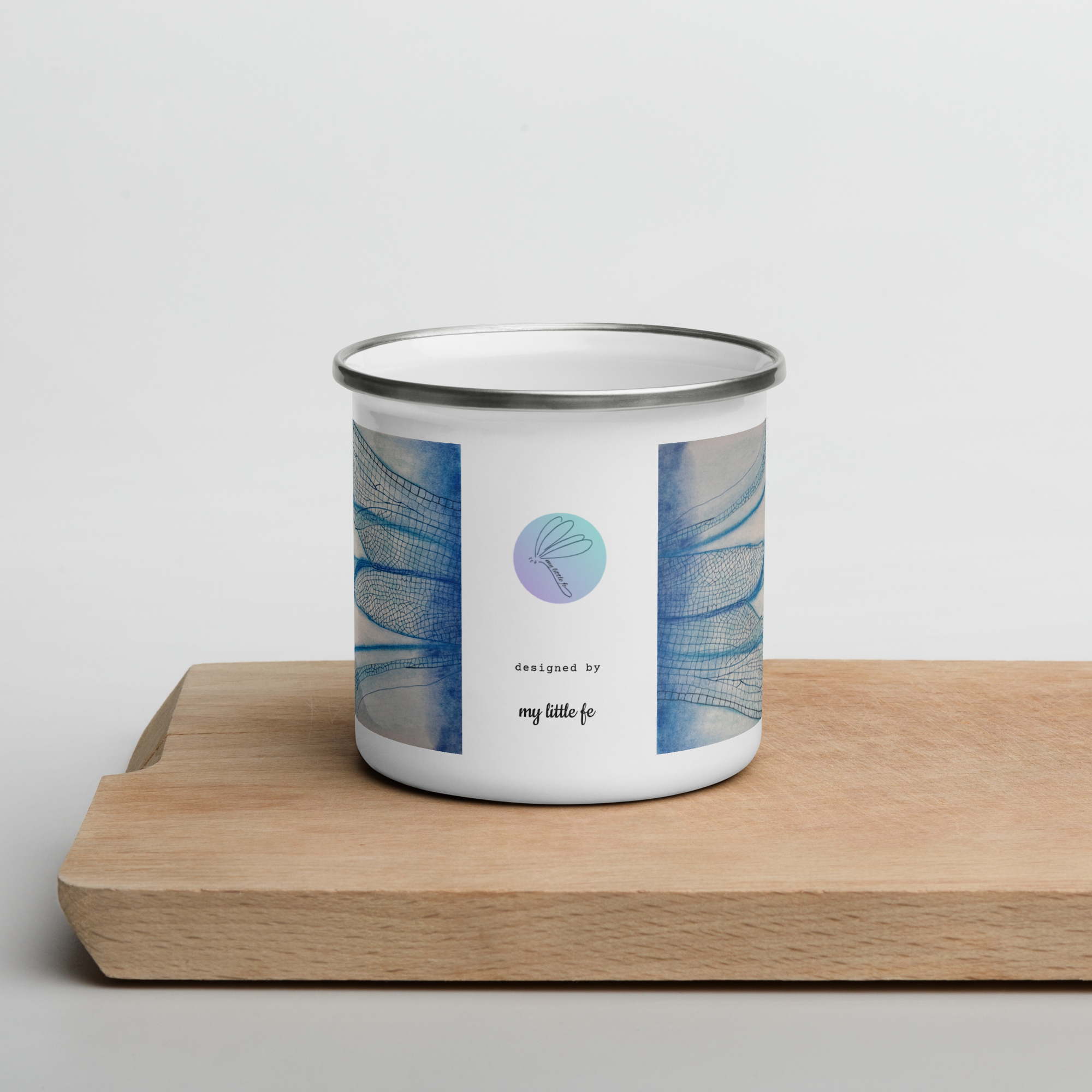 White enamel mug with blue dragonfly wings and a logo on a wooden surface