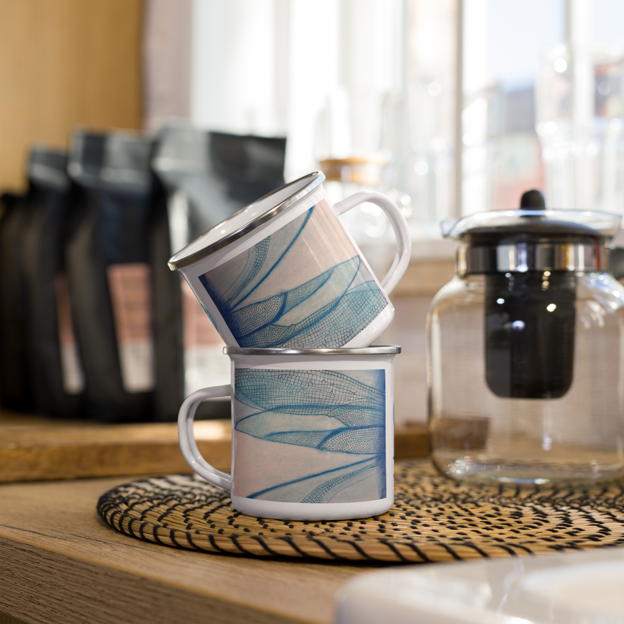 Two mugs with blue dragonfly wings designs stacked on a wooden surface with coffee-making equipment in the background.