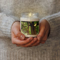 Hands holding a candle with a nature-themed design on a soft fabric background
